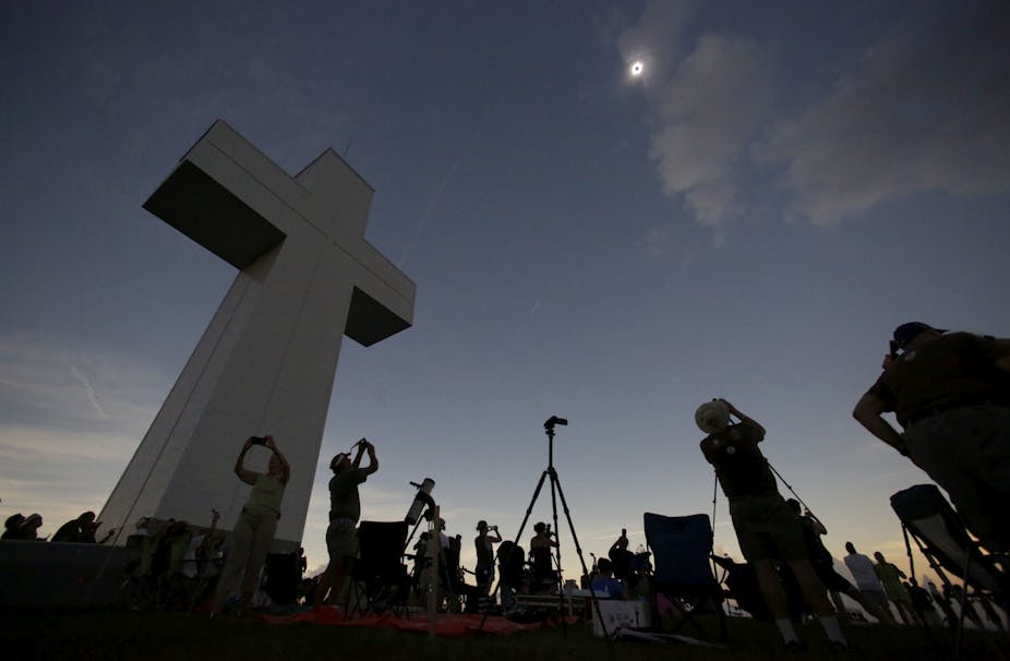 Several people standing near a large cross, while shielding their eyes and looking upwards at a solar eclipse.