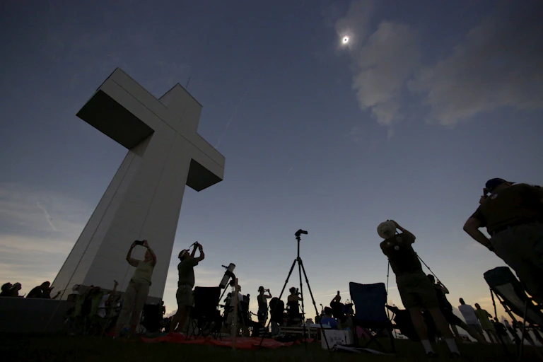Several people standing near a large cross, while shielding their eyes and looking upwards at a solar eclipse.