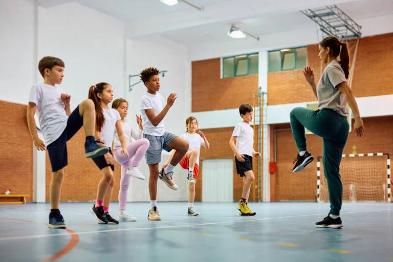 Dans un gymnase, une enseignante fait un mouvement d'échauffement en levant un genou. Face à elle, un groupe d'élèves filles et garçons reproduisent le même geste.