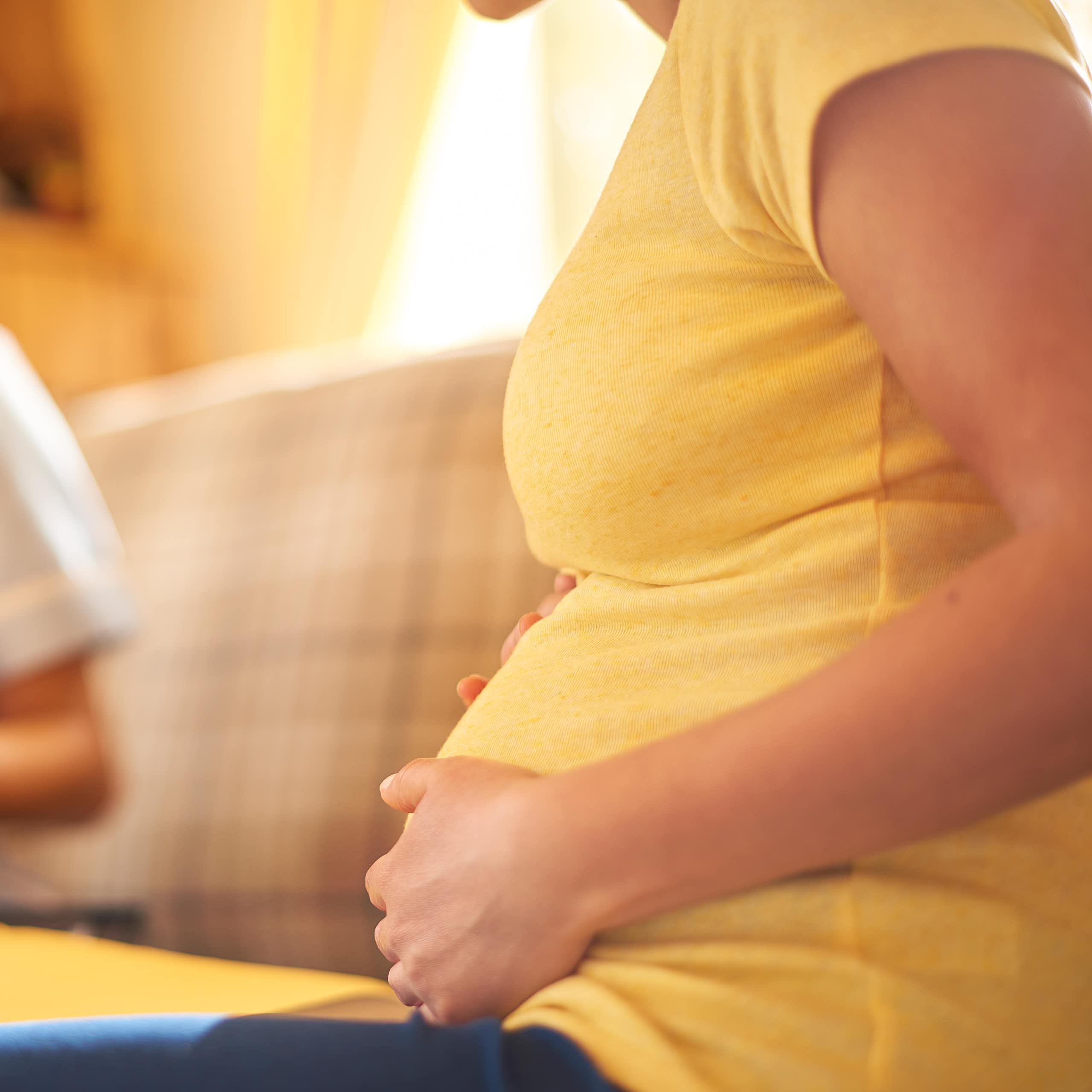 Pregnant woman holds her hands to her belly while sitting on a couch talking with a midwife who is taking notes.