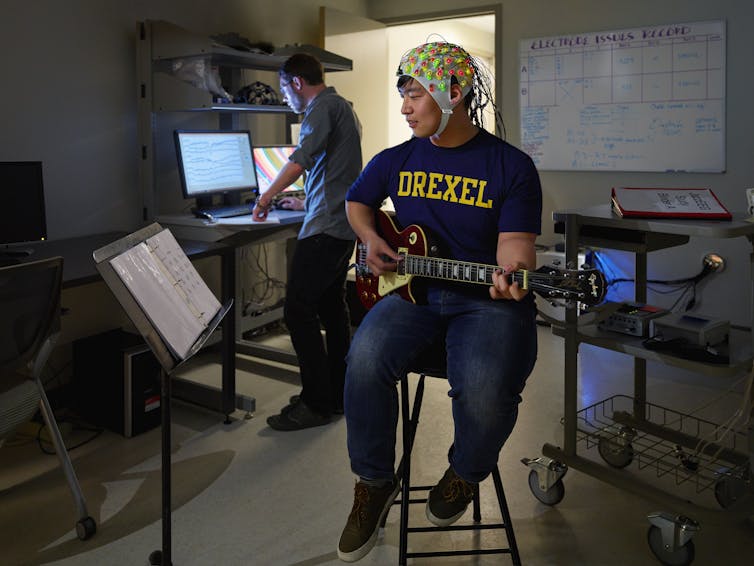 Young man plays guitar while wearing helmet covered in electrodes that measure brain activity