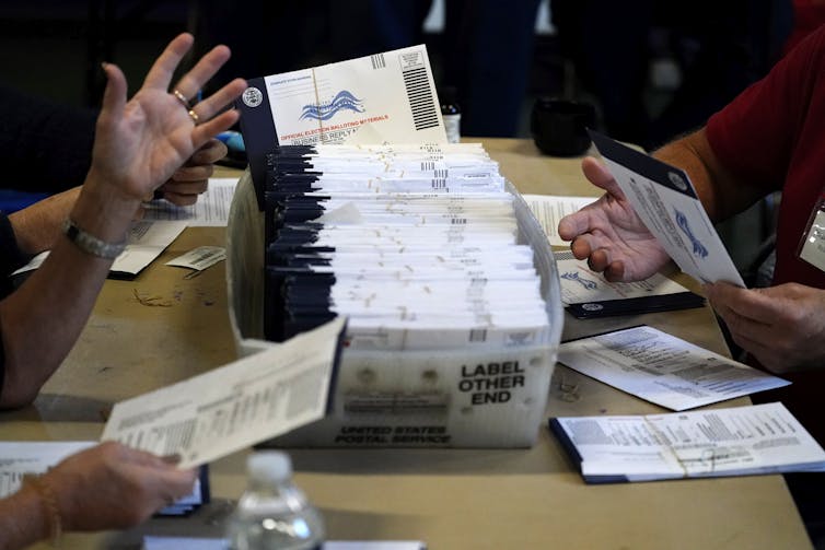 Una caja contiene muchos sobres electorales y las personas cercanas sostienen sobres individuales.