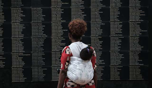A woman with a child in a sling on her back looks at a black wall with the names of people engraved on it.