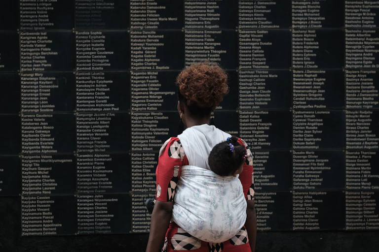 A woman with a child in a sling on her back looks at a black wall with the names of people engraved on it.