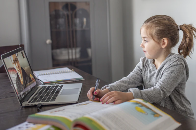 A young girl sits at a table with a pen and an open book while looking at a laptop.