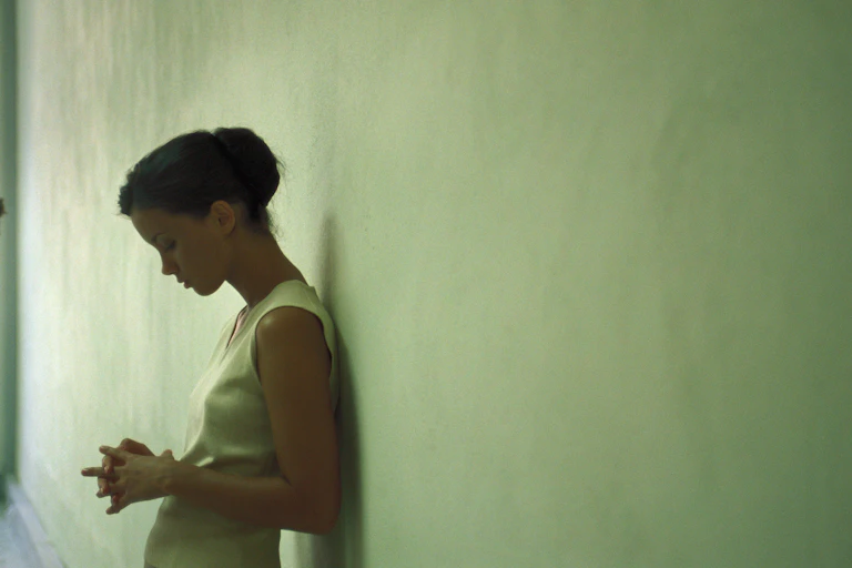 A young woman leaning against a wall looking pensive, eyes downcast.