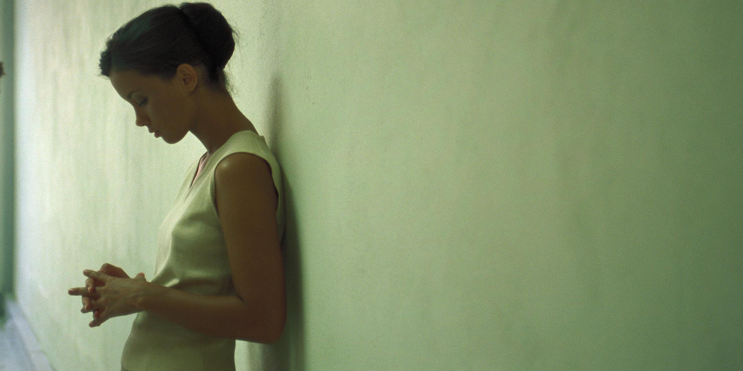 A young woman leaning against a wall looking pensive, eyes downcast.