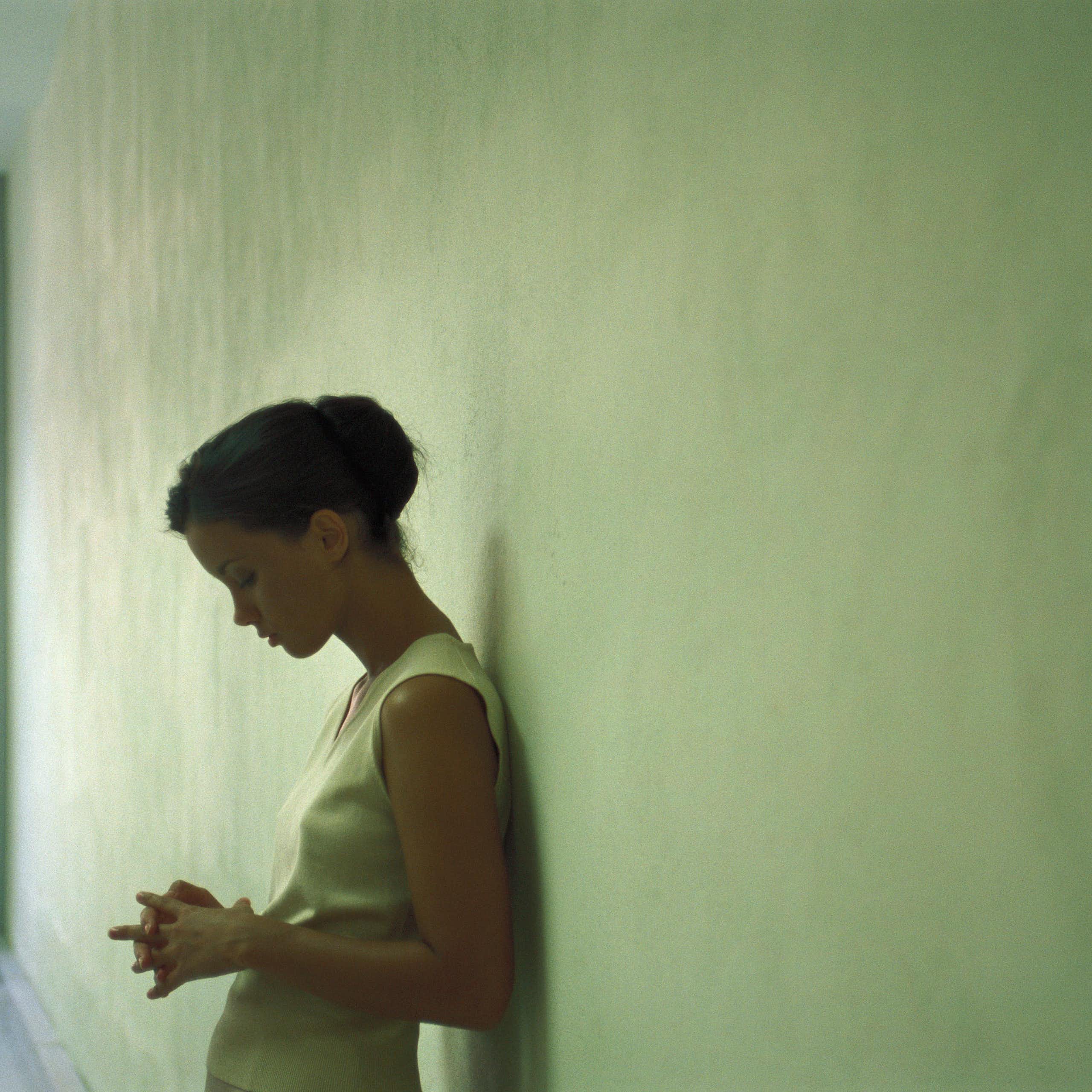 A young woman leaning against a wall looking pensive, eyes downcast.