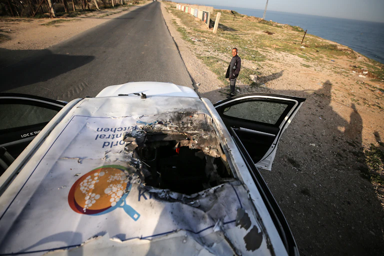 A man stands near a white vehicle that has a large hole in its top and its doors open. The road is empty and the ocean is nearby.