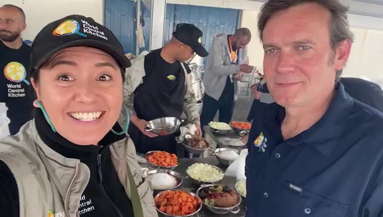 A woman smiles and stands next to a man, in front of a table filled with food. The woman wears a hat that says 'World Central Kitchen.'