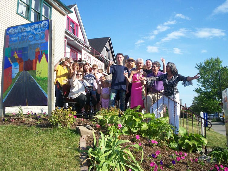 A group of people smile and pose, some lifting wine cups, in front of a garden.