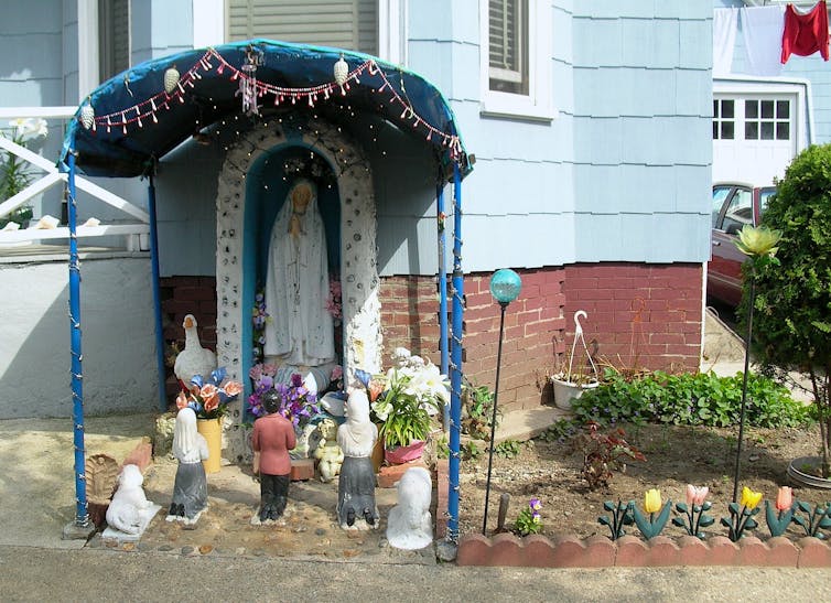 Religious shrine featuring Virgin Mary statue next to a home's front porch.
