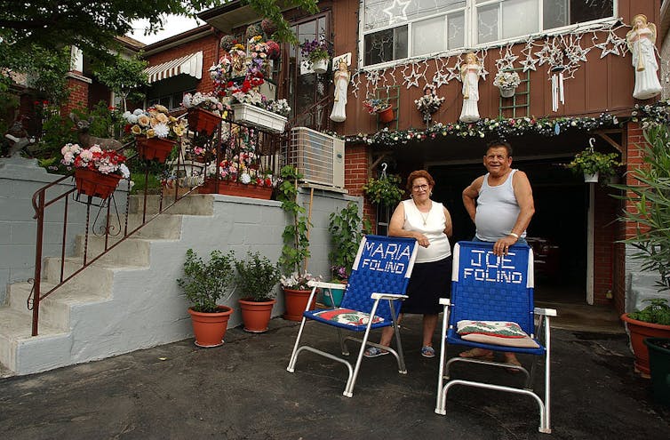 An elderly couple poses in their driveway in front of two chairs with each of their names written on them.