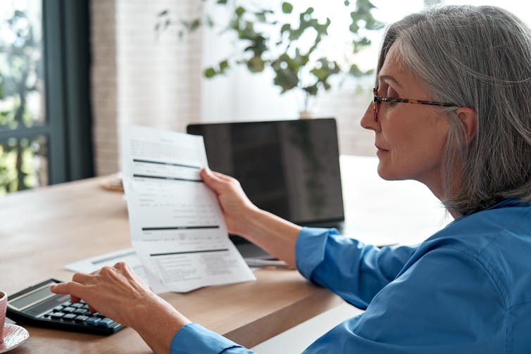 An older middle-aged woman with grey, shoulder-length hair presses buttons on a calculator while holding a paper bill in one hand