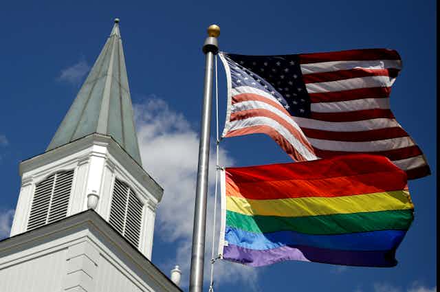 A white church steeple next two an American flag flying on top of a rainbow striped flag.