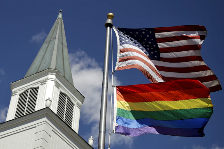 A white church steeple next two an American flag flying on top of a rainbow striped flag.