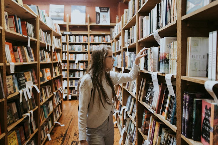 A young woman picks a book from a shelf in a bookshop.