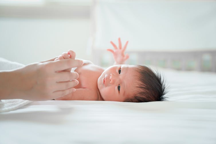 A baby lying on a bed, with an adult holding its hand.