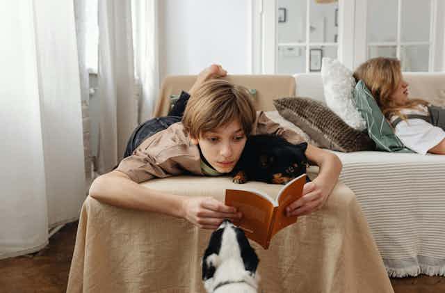 A teenage boy lies on a couch with a dog, reading a book.