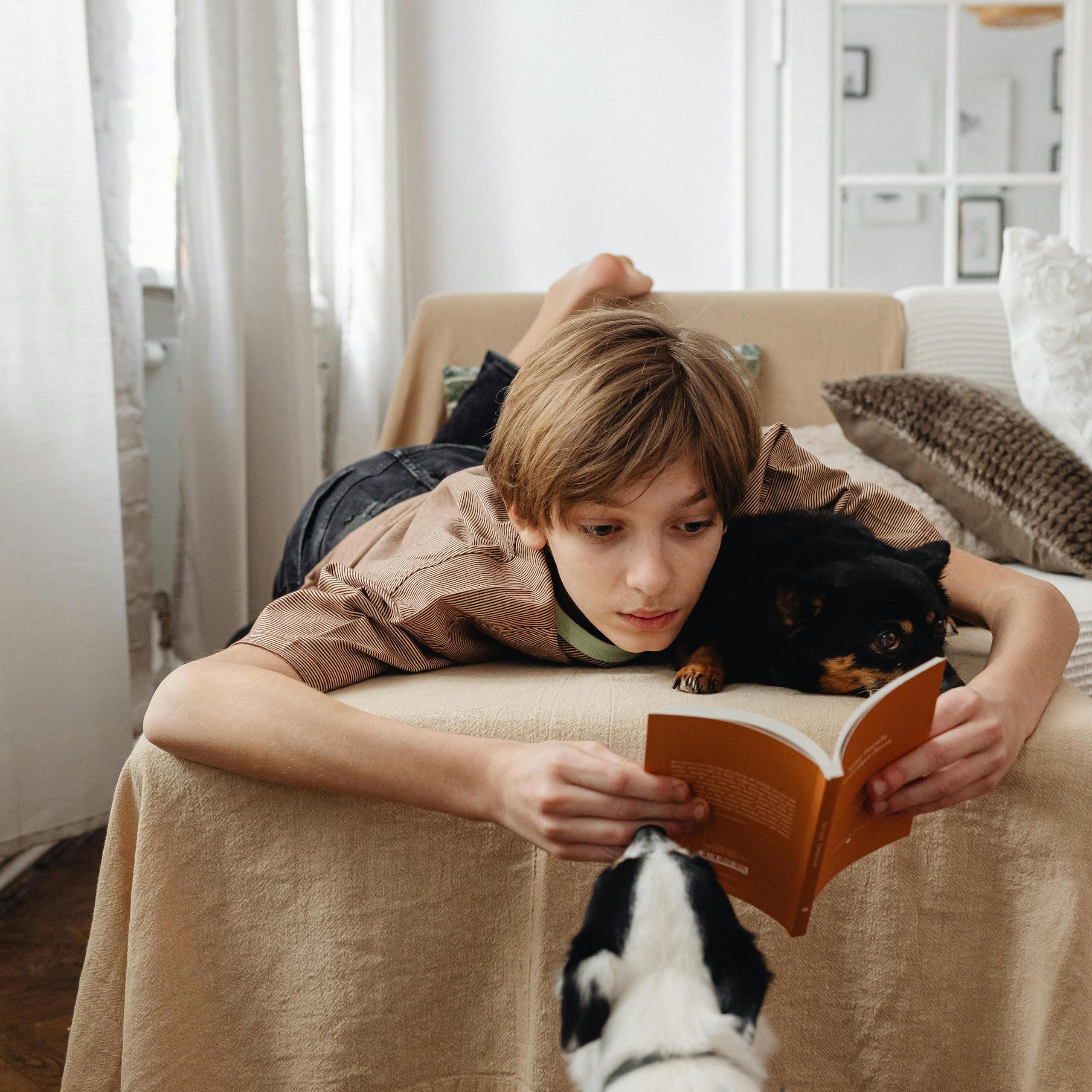 A teenage boy lies on a couch with a dog, reading a book.