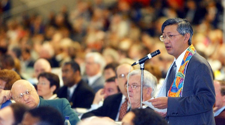 A man in a suit and glasses speaks into a microphone in front of a crowded room of seated people.