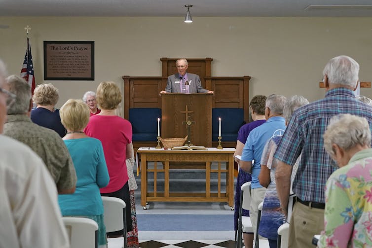 A man in a suit speaks at a pulpit in front of a room of standing people, most of whom are older.