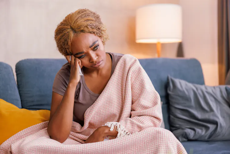 A sad woman sits on the couch with a blanket covering her body.