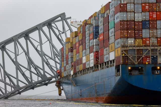 A cargo ship carrying stacks of boxes crashes into a metal structure. 