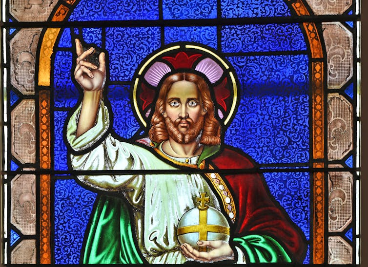 A stained glass image of Jesus, with a halo around his head, raising the fingers of one hand towards the sky while holding an orb in the other.