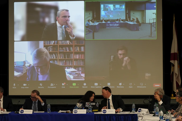 People at a long table with name placards, with a large screen featuring people on it behind them.