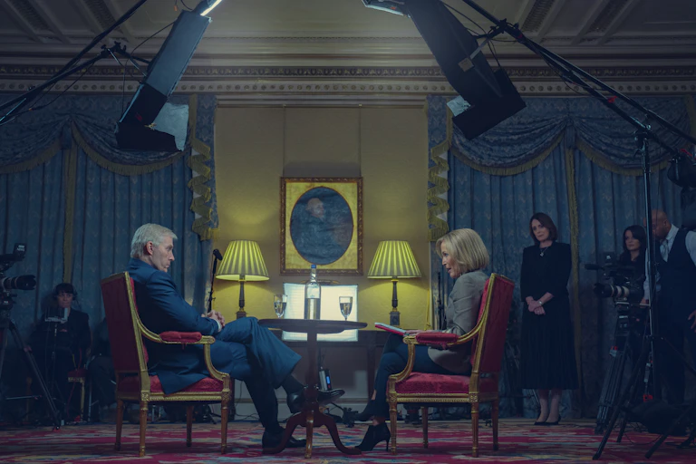 Still image of Sewell and Anderson in character, seated in red chairs doing the infamous Newsnight interview beneath tv cameras and lights