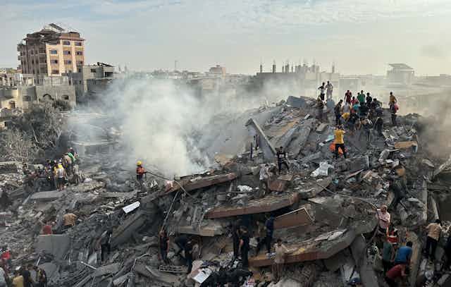 Workers and residents stand on a pile of rubble with one building standing in the background.