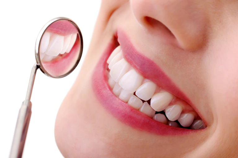 A smiling woman holds a dental mirror up to her bright white teeth.