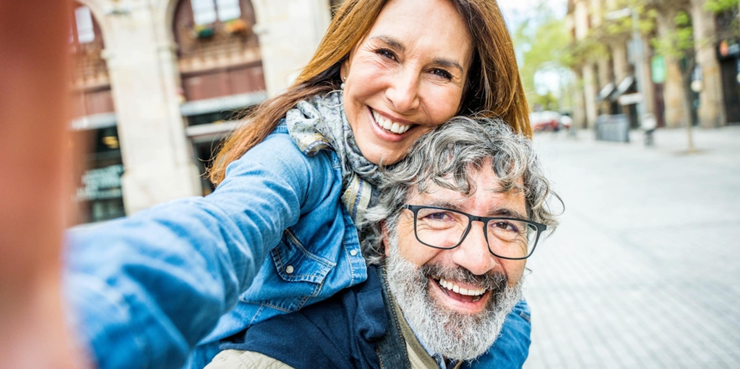 Older happy couple, woman on man's back, taking a selfie in a piazza or town square