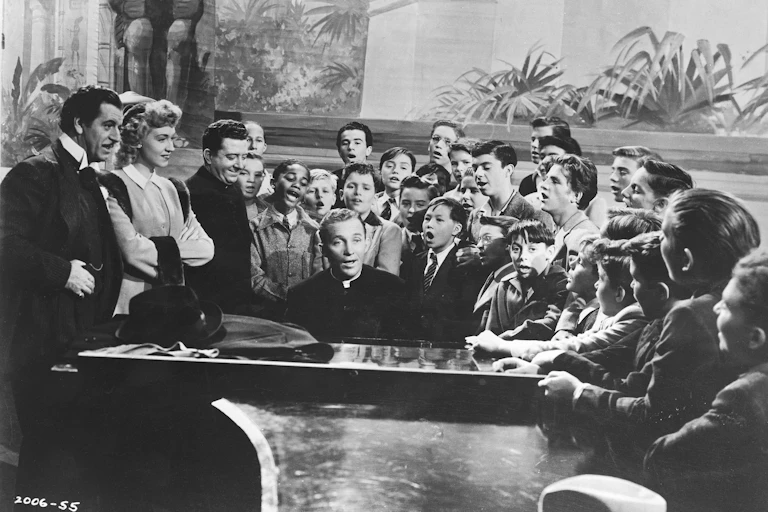 Un cura dirige desde el piano una sesión de canto para niños mientras algunos adultos observan.