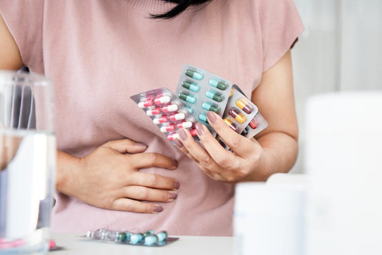 A woman holds multiple packs of antibiotics with one hand, and presses her other hand onto her stomach in pain.