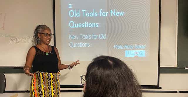 Foto mostra professora Rosy Mary em sala de aula. No projetor, lê-se Old tools for new questions