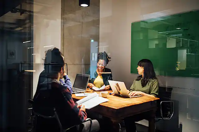 Businesspeople speak in meeting while working late seen through glass door at office.