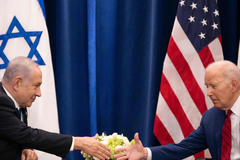 Two men sit facing each other and extend their hands toward each other. They sit in front of Israeli and U.S. flags.