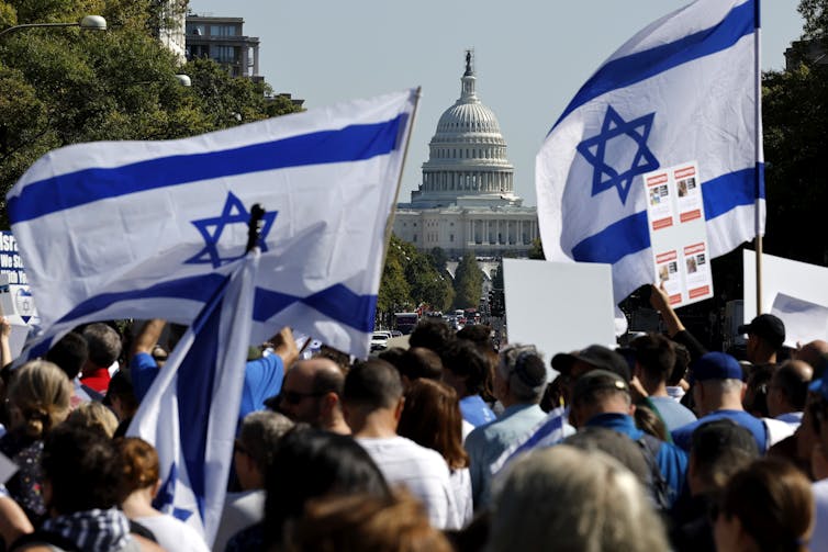 A large crowd of people stand near the US Capitol and hold Israeli flags that are white with blue stars.