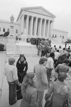 A black-and-white photo shows a line of people winding their way up the steps of the neoclassical U.S. Supreme Court building.