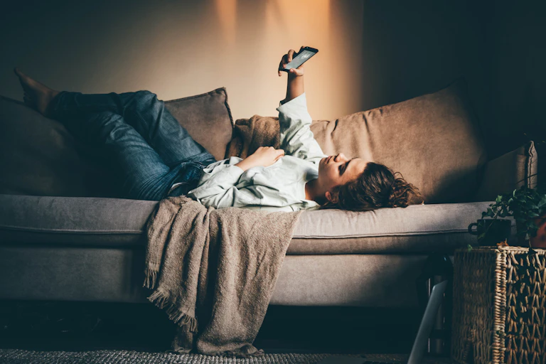 A young woman lying on a sofa at home scrolling on her phone, a laptop is sitting on the floor