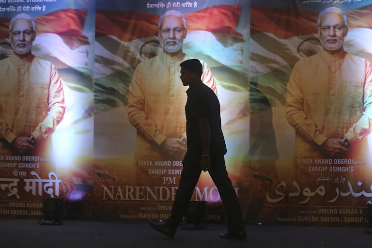 A silhouette of a man walking past three identical posters showing a gray-haired man, dressed in a long saffron-color shirt, with the flag of India in the background.