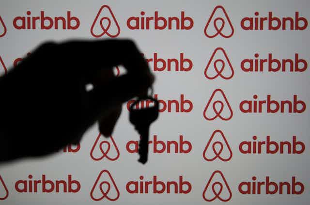 A hand is seen holding a key in silhouette before a computer screen displaying the Airbnb logo