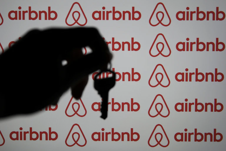 A hand is seen holding a key in silhouette before a computer screen displaying the Airbnb logo