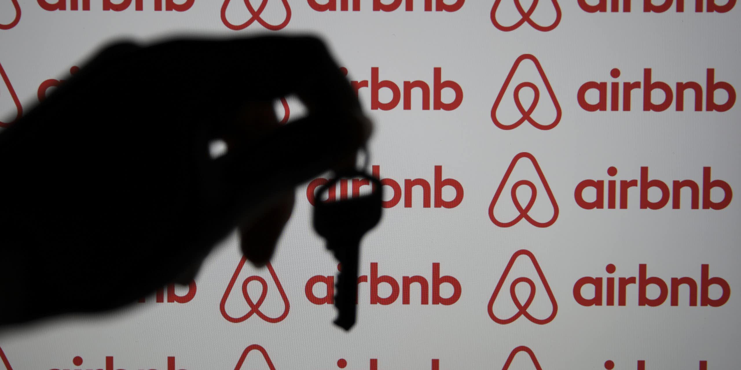 A hand is seen holding a key in silhouette before a computer screen displaying the Airbnb logo
