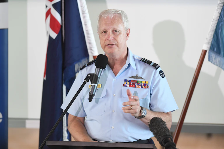 A man in a blue shirt with military badges delivers a speech behind a lectern