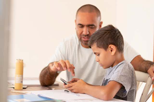 A man and a boy sit at t table with pencils, books and a calculator. 