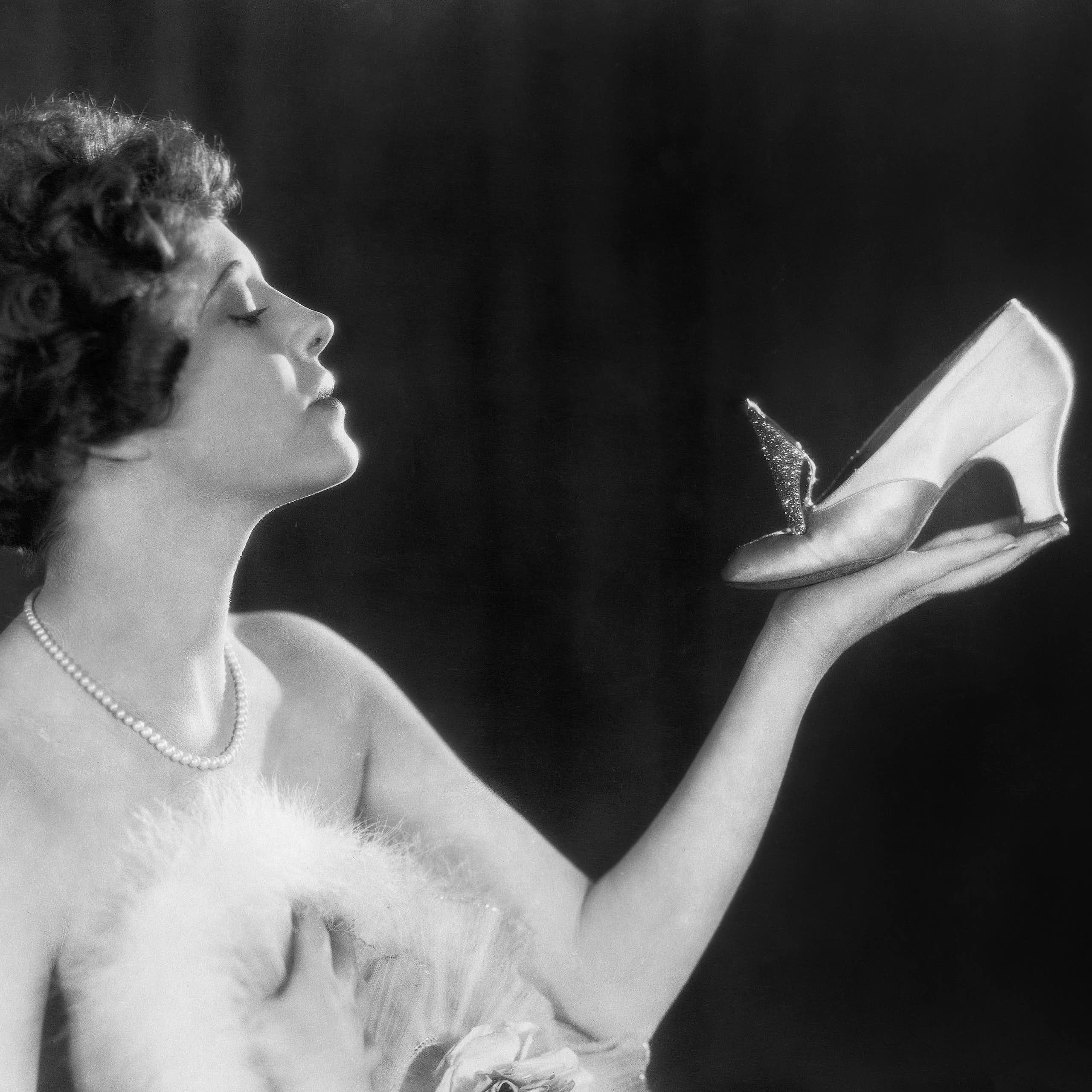 A 1940s woman holds a heeled shoe in one hand while looking at it