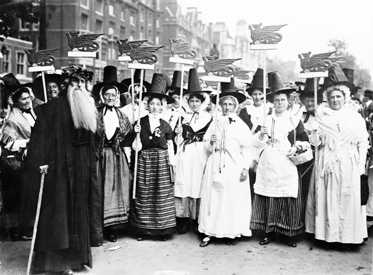 A black and white photo of a group of women in traditional Welsh costume with tall black hats hold models of dragons above their heads. A bearded man stands next to them.